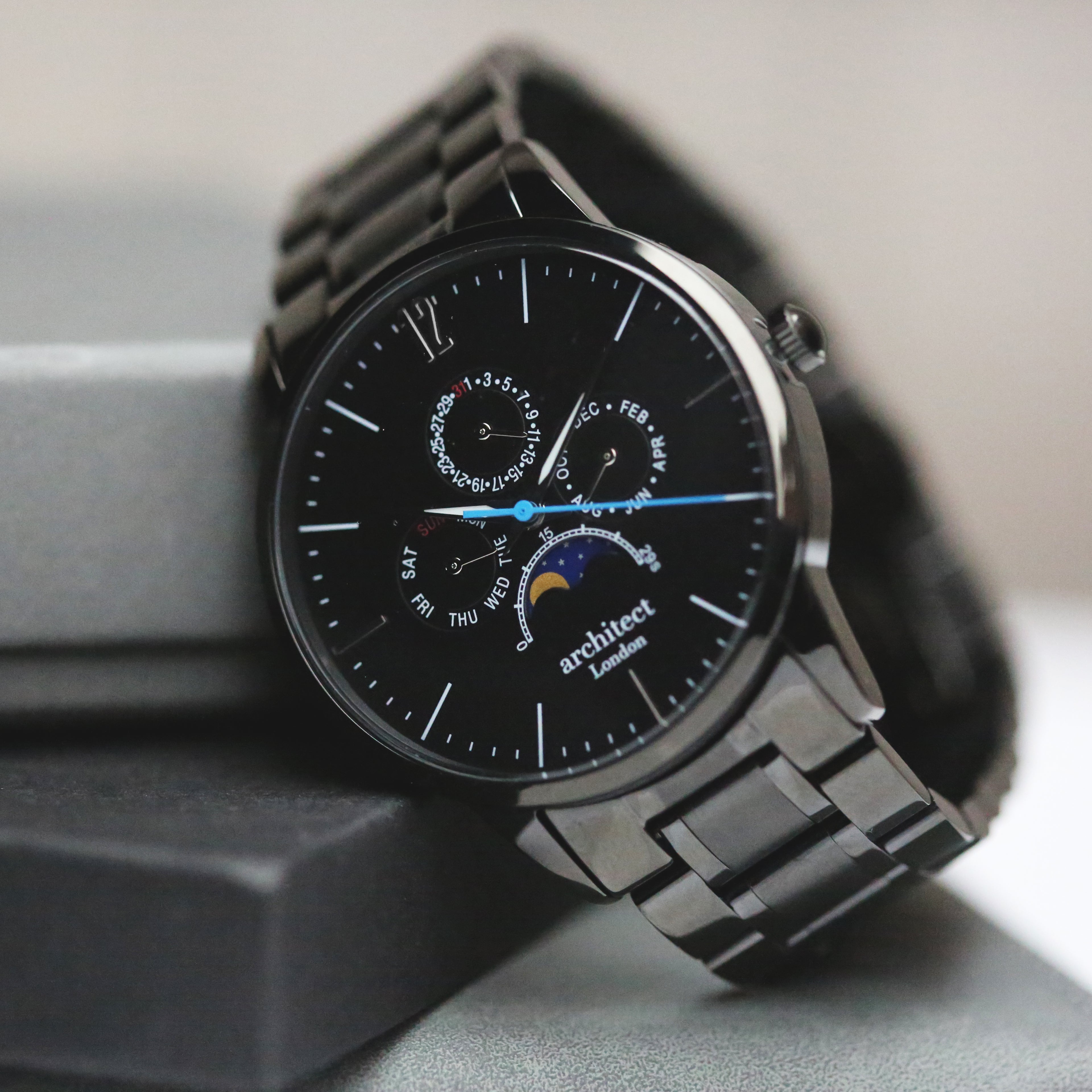 Sleek black Architect London watch with moonphase display and polished stainless steel bracelet on grey box.