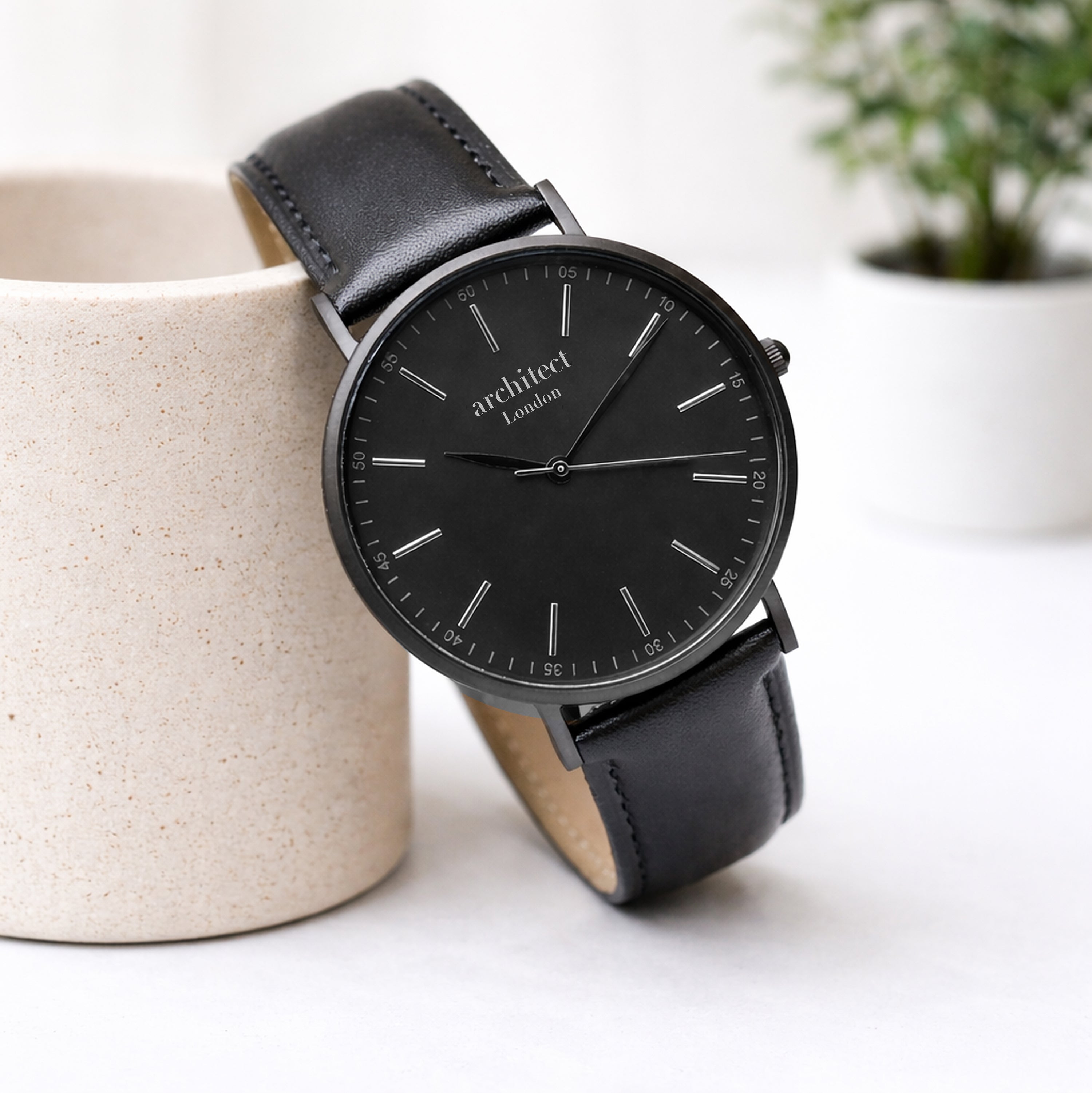 Black wedding watch with leather strap on a white surface with a blurred plant in the background