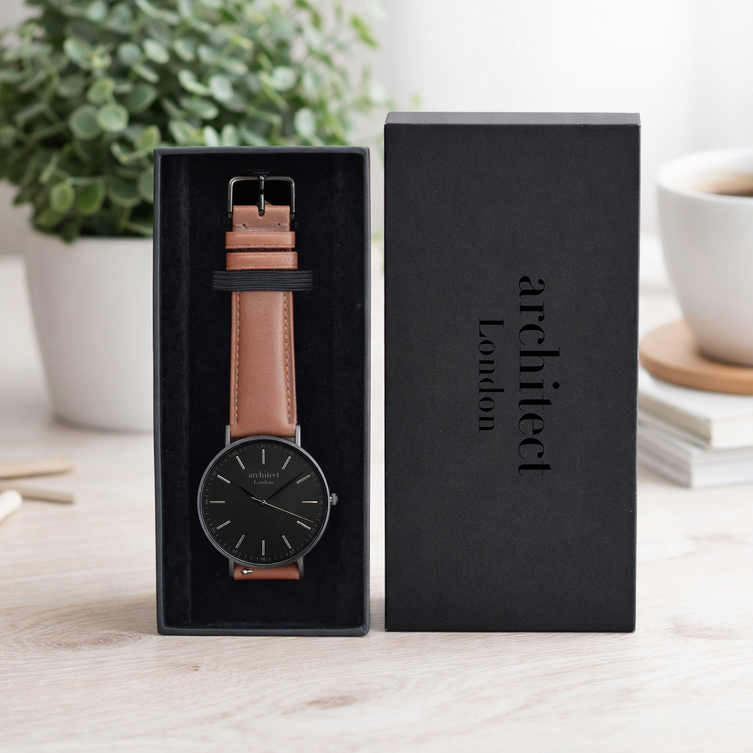 Watch with brown leather strap in a black box, branded 'architect london', on a light wooden surface.