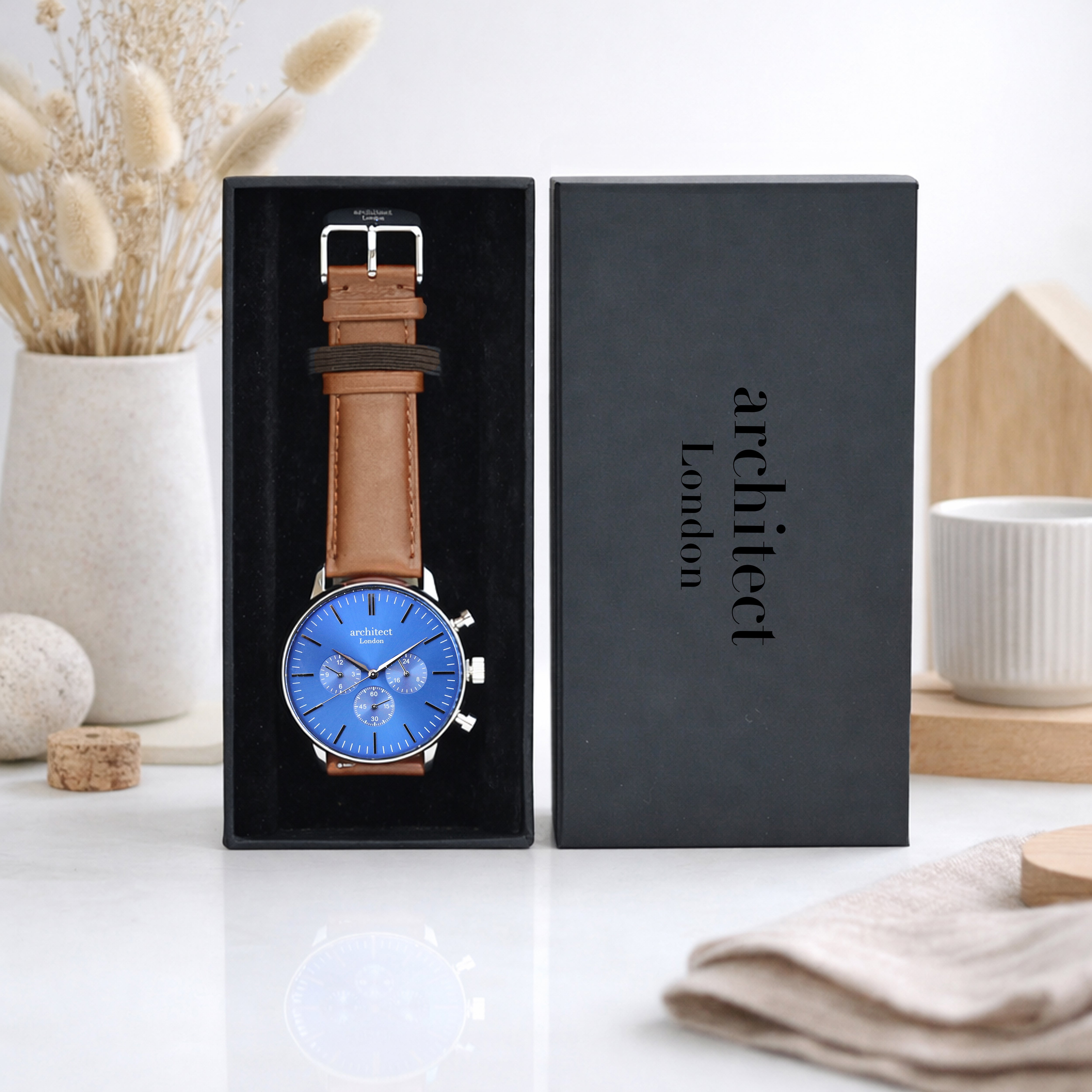 Watch with brown leather strap and blue face in a black box, branded 'Architect London', on a white surface with decorative items.