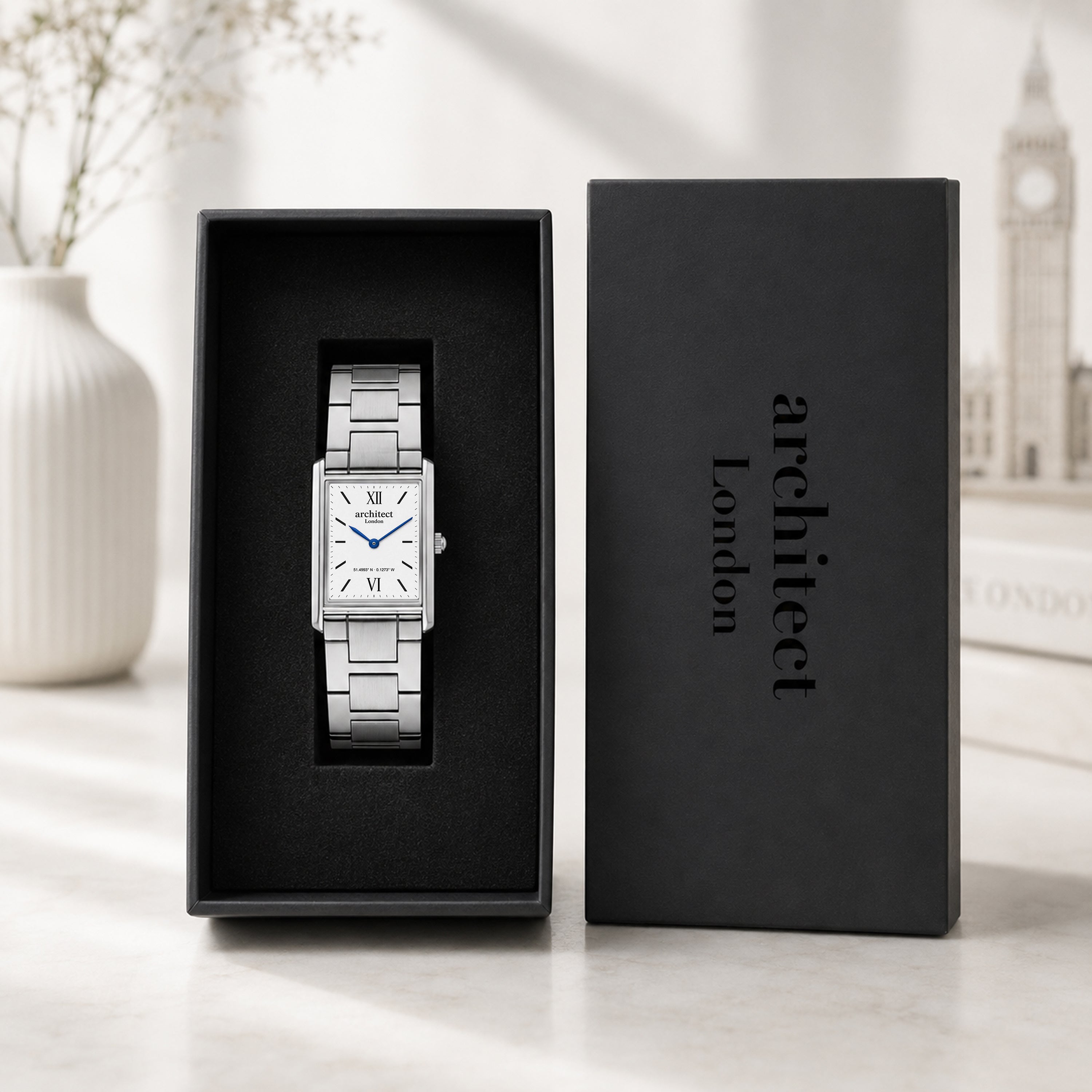 A silver rectangular analog watch with a white dial and blue hands is displayed inside an open black presentation box next to its matching black lid, which is printed with the brand name 'architect London'. The scene is set on a light marble surface with a blurred white vase and a faint Big Ben background.