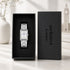 A silver rectangular analog watch with a white dial and blue hands is displayed inside an open black presentation box next to its matching black lid, which is printed with the brand name 'architect London'. The scene is set on a light marble surface with a blurred white vase and a faint Big Ben background.